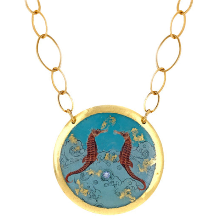 A gold leaf over brass disc pendant depicting two seahorses with a blue ocean background, attached to a gold-plated chain.